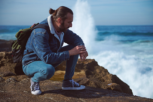 Bearded young adult male traveler checking time on his wristwatch, or using compass for searching the north direction, sitting on the edge of a rocky cliff against huge waves breaking on the headland