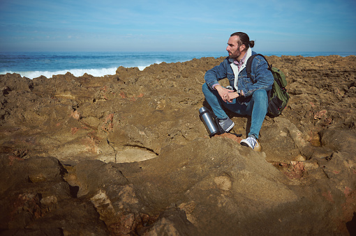 Relaxed bearded male traveler, adventurer, backpacked tourist sitting on a rocky cliff, looking into the distance, enjoying the view of Atlantic ocean at sunset. Beautiful waves crushing on headland