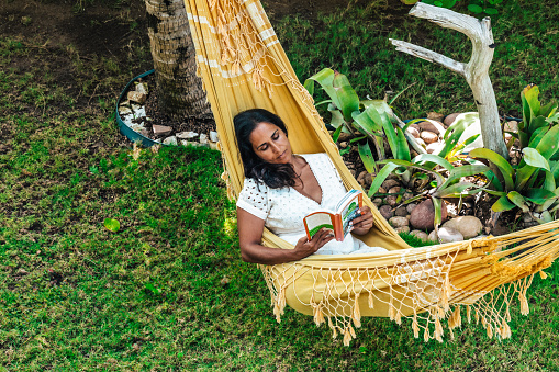 view on mid adult brazilian woman reading book in hammock in the garden in summer