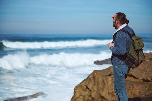 Absorbed man traveling alone, carrying a backpack, looking into the distance, watching horizon, enjoying the view of beautiful nature with splashing waves on the headland, standing alone on the cliff
