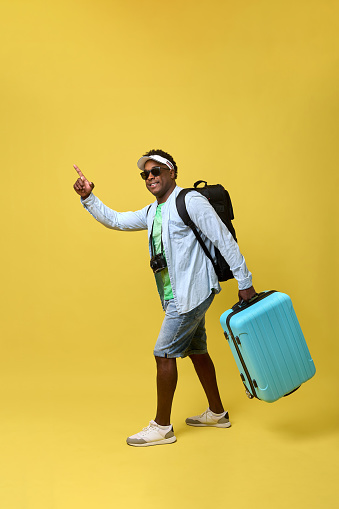 Cheerful young African man dressed in summer clothes walks with a suitcase and backpack and points his finger at the pointer. Young man in shorts and shirt walking with luggage on yellow background