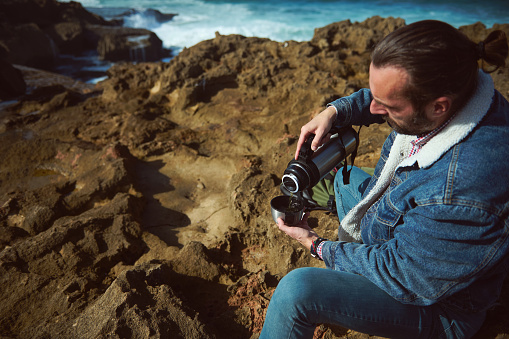 Caucasian bearded young man tourist traveler in casual denim, sitting on the rock by sea, enjoying his coffee break in the nature, pouring some hot dink from a stainless steel thermos flag into a mug