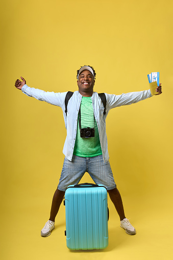 Portrait of a happy adult black man with hands on a yellow background in a shirt and denim shorts. An African smiles, travels and relaxes with a comfortable suitcase and backpack