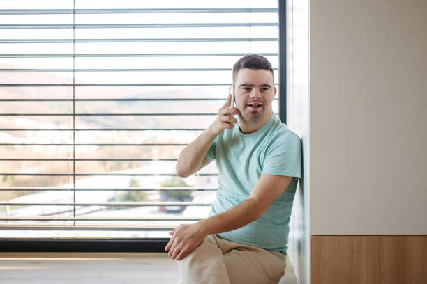 istock Young man with Down syndrome sitting by window, making phonecall with smartphone. 2015396664