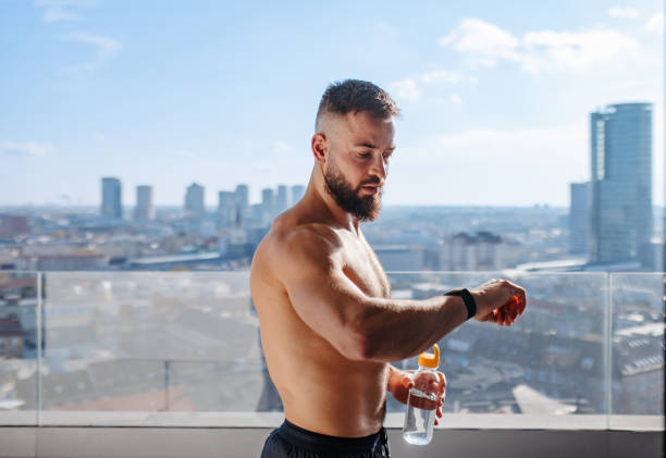 Fashion handsome healthy muscular man with sunglasses in casual outfit lifted his shirt and looks at his abs near a white wall with sunlight. Morning and diet