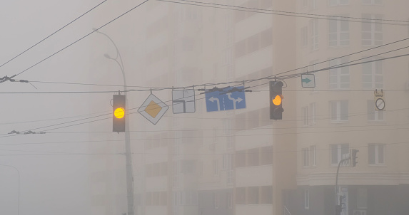 Blinking yellow traffic lights at an intersection in the city, with a high-rise apartment building in the background. Fog, limited visibility, trolleybus wires. High quality photo