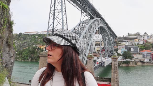 Stylish woman in glasses and checkered hat stands in front of Ponte Luis I, Porto
