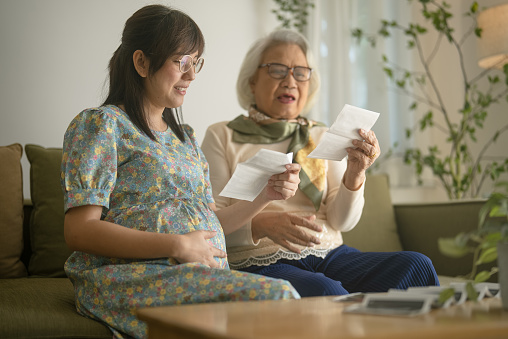 Senior grandmother and young pregnant daughter watching pictures in ultrasound scan, smiling and speaking while sitting on sofa in the evening.
