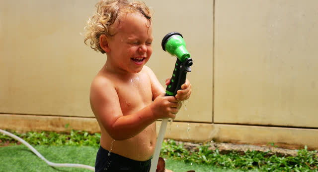 Small child pointing handheld sprinkler at his face is surprised by weak stream