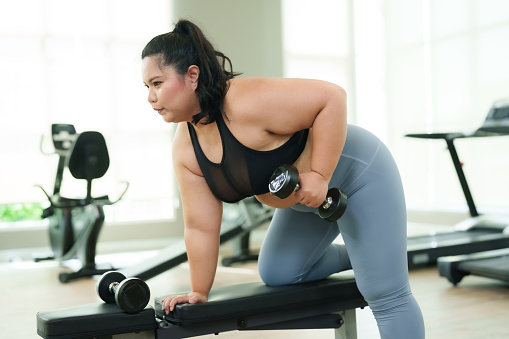 Plus-size Asian woman exercise in gym, activewear engaged in a dumbbell workout on bench, concentrating on fitness regimen. Focused female performing dumbbell rows, dedication to workout routine