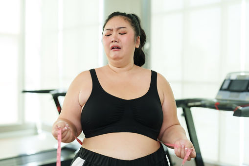 Chubby Asian woman exercise in gym, visibly upset while measuring waist, moment of struggle on fitness journey, Emotionally overwhelmed with measuring tape, confronting weight loss challenges