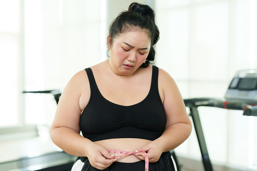 Plus-size Asian woman exercise in gym, visibly upset while measuring waist, moment of struggle on fitness journey, Emotionally overwhelmed with measuring tape, confronting weight loss challenges