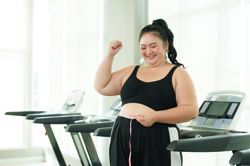 Plus-size Asian woman exercise in gym, Raise hands in joy progress with tape measure, triumph in health and fitness journey. Elated female measuring waist, a victorious moment in her workout regimen