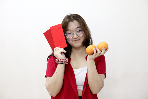 Portrait shot of an Asian teenage girl holding mandarin oranges and red envelopes (hongbao) and smiling at the camera