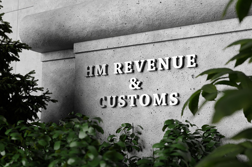 Silver sign of Home Office Revenue and Customs on a concrete wall with green plants as foreground. Illustration of the concept of taxation