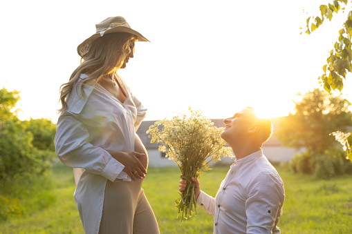 A man in a hat kneels on the grassland in front of a pregnant woman, offering a bouquet of flowers. Both appear happy and surrounded by nature