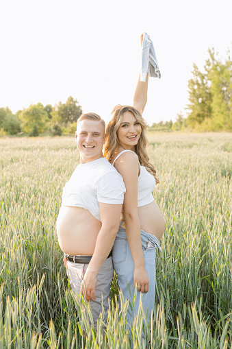 A man and a pregnant woman are smiling and posing in a field with natural landscape, under the sunlight. They are wearing shorts and surrounded by grass, with the sky as background