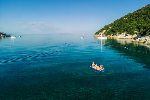 Amazing coast with beautiful beach on Ionian sea and three women enjoying while sitting together and paddling on stand up paddleboard. October in Greece, off season. Filiatro beach, Ithaca island, Greece