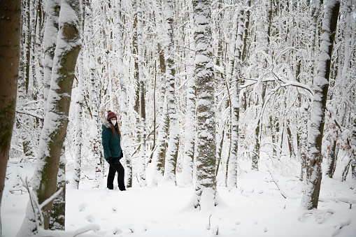 A lone figure stands in a forest blanketed by snow, with trees cloaked in white extending into the background. The person is wearing a teal jacket and a beanie, and a white mask covers their face, highlighting the solitude of the winter setting