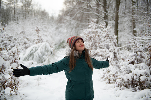 The photo captures a smiling woman with outstretched arms, enjoying the snowy environment. She wears a teal winter coat and a colorful hat, surrounded by snow-covered shrubs and trees in a dense forest setting