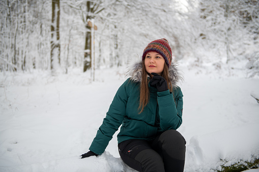 An image featuring a woman seated on a snow-covered ground in a forest, looking thoughtful. She is dressed in a teal winter jacket and a multicolored knit hat, with her hand resting on her chin, surrounded by the white expanse of winter