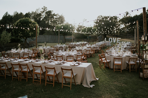 Very general photograph of an outdoor wedding banquet. Different tables, long and round decorated. Nature environment, trees and grass, illuminated with garlands. At one end a luminous sign with the word love.