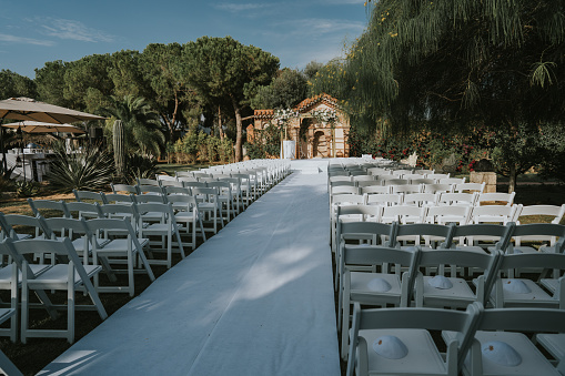 General shot of a Jewish ceremony before it begins. We see a multitude of white wooden chairs, placed in two rows, separated by a white carpet. On top of the chairs is a white yarmulke with gold embroidery.