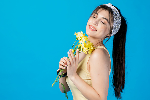 Side vie of slim, brunette lady with hair band and ponytail posing indoors, holding yellow flower, smiling. Concept of beauty.