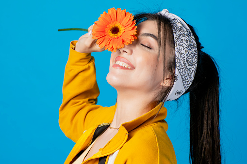 Side view of pretty, young woman posing with orange flower, raising hand, touching face, smiling. Isolated on blue studio background.