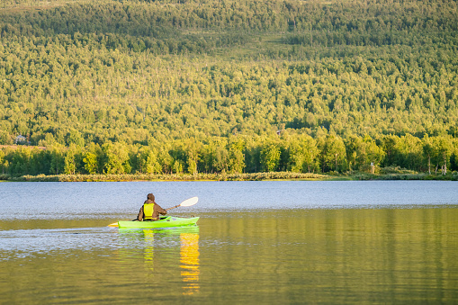 Mature women in green safety life jacket kayaking in green kayak. Rear long distance photo on still water with blurry green mountain forest background. Sweden.