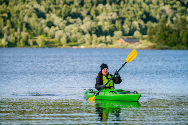 зрелые женщины в зеленых спасательных жилетах катаются на каяке в зеленом каяке, она смотрит в камеру. фотография спереди на стоячей воде н� - women kayaking life jacket kayak стоковые фото и изображения