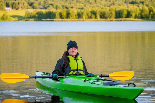 Mature women in green safety life jacket sits in green kayak and look at camera holding yellow paddle across boat. Very close up photo on still water with blurry background. Sweden