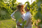 A happy pregnant woman wearing a hat stands next to a man in a vineyard