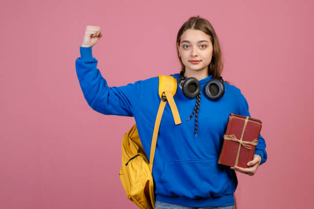 Front view of young student standing, raising hand, showing strong. Pretty schoolgirl wearing casual clothes, looking at camera, holding bunch of old books. Concept of youth.