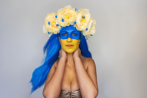 Creative close-up portrait of young Ukrainian patriot woman with yellow blue face art, wreath of flowers on head on isolated gray background. Flag Day, Independence Day in Ukraine. Copy space