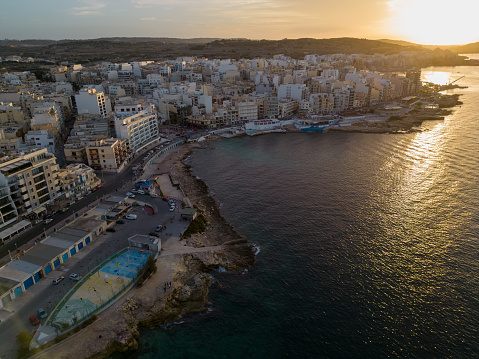 St. Paul's Bay with playground by the sea at sunset. Aerial view towards Bugibba square and the golden sun with reflection on the Mediterranean sea. Generic residential and commercial buildings.