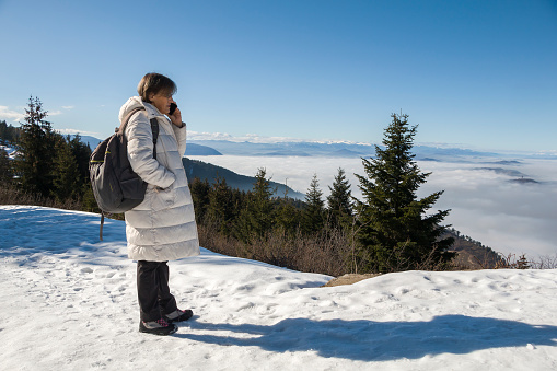 An elderly lady in warm clothes communicates on the phone. View from the mountain to the polluted city below which is in the fog