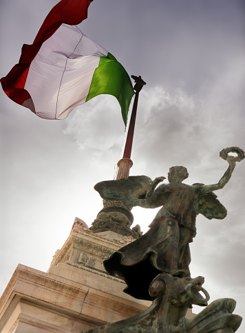 Rome, Italy: the Vittoriano. The italian flag flying under a grey sky under a statue at the Altare della Patria (altar of the homeland), a national monument built (1885-1935) to honor King Vittorio Emanuele II