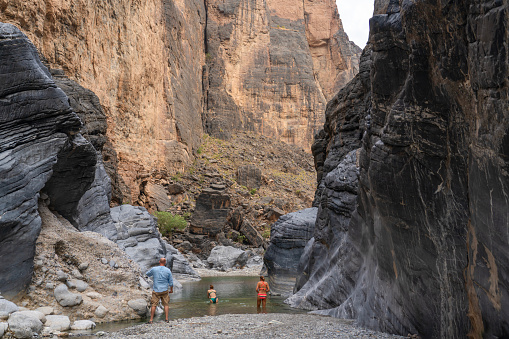 Two women swimming in Snake canyon, one men watching over them on the river bank in Wadi Bani Awf—one of Oman’s most picturesque valley.