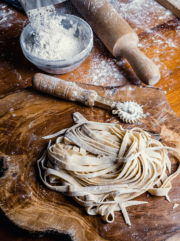 Working with handmade pasta. Close-up on a type of handmade pasta called fettuccine, left to rest on a wooden cutting board. Some useful tools for working: a rolling pin, a pastry wheel cutter and some flour to add to keep the consistency of the finished product soft.