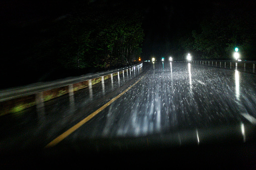 A driver's point of view looking ahead at some hilly curves through a speeding car's windshield late at night during a non-stop extreme weather rain storm. The car's headlight high beams are battling with torrential streaks and splashes of seriously intense downpour and causing road reflectors and other road signs ahead to blaze brightly with lens flare illumination. No need to use windshield wipers because the continuously heavy rain provided a constant clear stream of water flowing across the glass.