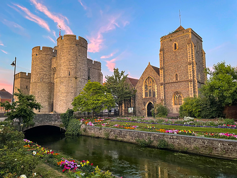 Colorful sunset clouds over medieval Westgate Towers and Guildhall in Canterbury. Amazing view of blooming flowers on the banks of River Stour that flows slowly through picturesque historic town.