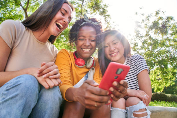 istock Group joyful friends using mobile sitting outdoor. Three laughing multiracial women scrolling phone 2016281804