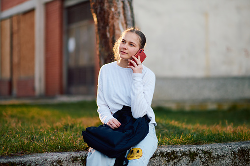 A beautiful blonde teenage girl sits on a park bench, using her smartphone and wireless headphones to prepare for school, with her school backpack beside her on the bench.