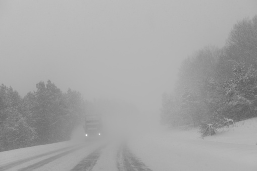 front passenger view of a highway in a blizzard, with fairly heavy traffic, a truck coming from the opposite direction, fog and limited sight, trees on the sides, snow on the road.
