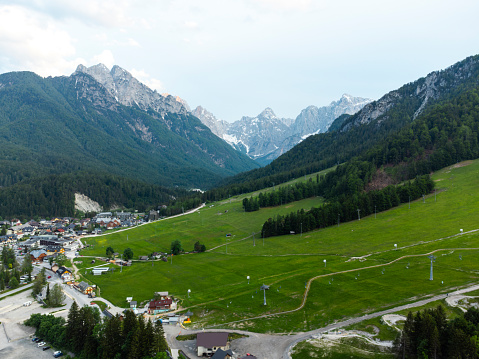 Aerial view of a lush valley with a small town, surrounded by majestic mountains and green forests under a cloudy sky.