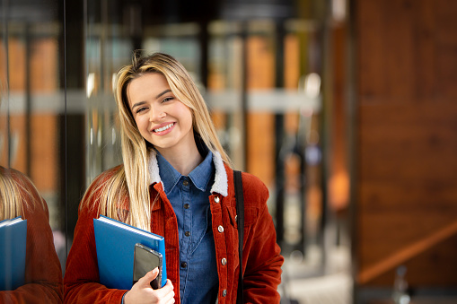 A confident young adult girl student stands in front of a glass modern building, holding a backpack, books, and phone. With a smile, she faces the camera, symbolizing readiness for digital learning. Ideal for educational promotions or content showcasing modern student life.