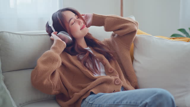 Asian woman relaxing on comfortable couch with wearing headphones in living room. Female enjoys listening chill music audio sound feeling no stress at home. slow motion