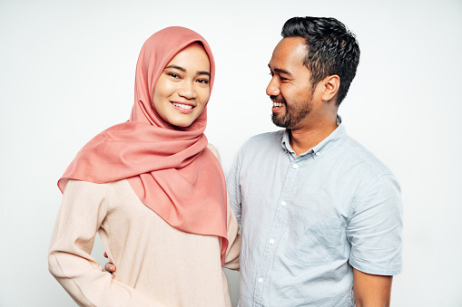 Young happy Malaysian couple standing together side by side. Young man looking over to the women with a smile. Bright smiling Malaysian woman with headscarf looking towards the camera. White Background Portrait Studio Shot. Kuala Lumpur, Malaysia.