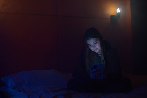 Girl in the twilight sits on the bed scrolling the news on her phone